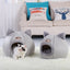 Two gray pet houses with cat ears on a white carpet, with a cat peeking out from one of them. Behind them is a beige sofa with colorful cushions.