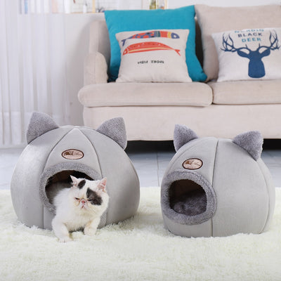 Two gray pet houses with cat ears on a white carpet, with a cat peeking out from one of them. Behind them is a beige sofa with colorful cushions.