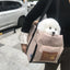 Small white dog in a beige bag with text, held by a person on a sidewalk.