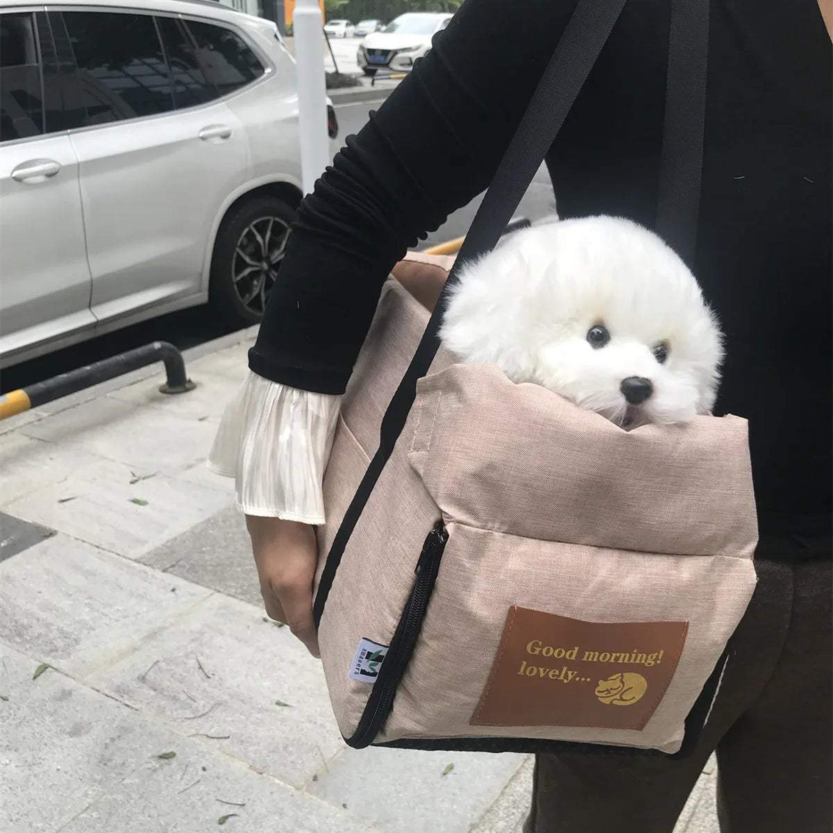 Small white dog in a beige bag with text, held by a person on a sidewalk.